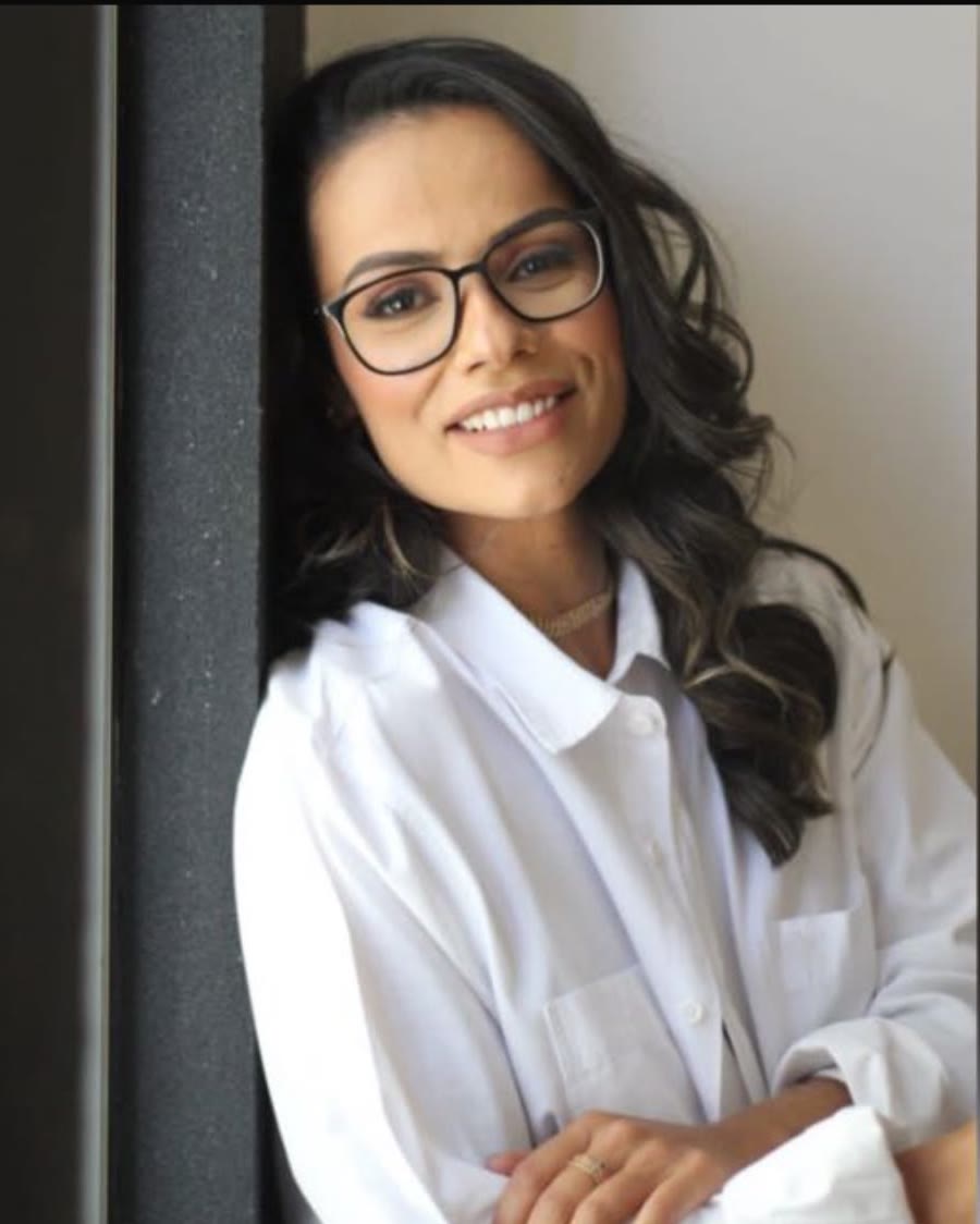 Dra. Cecília Ribeiro sorrindo, de óculos e camisa branca, encostada no batente de uma porta